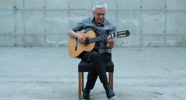 Caetano Veloso playing an acoustic guitar whilst on a chair