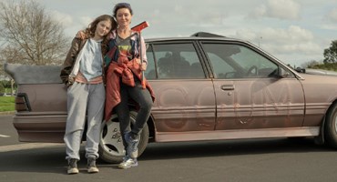 A teenage girl and a woman are smiling and leaning against a car with Scum written on it